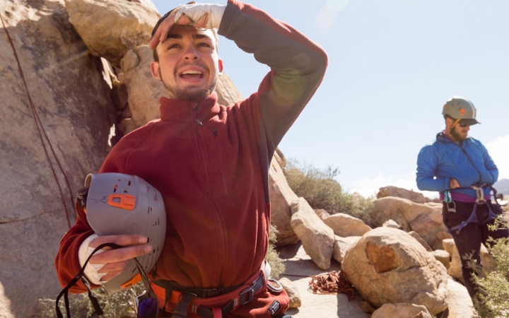 two students in rock climbing gear stand amongst rocks and green shrubs
